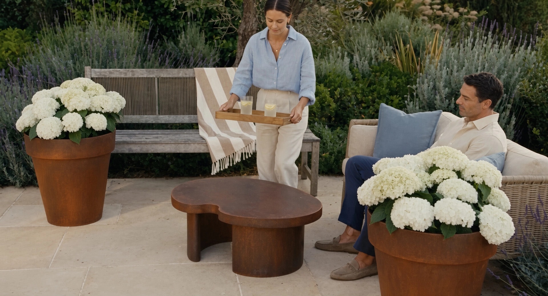 Pots de fleurs coniques Calypso en acier Corten aux hortensias blancs, une femme pose un plateau sur une table basse