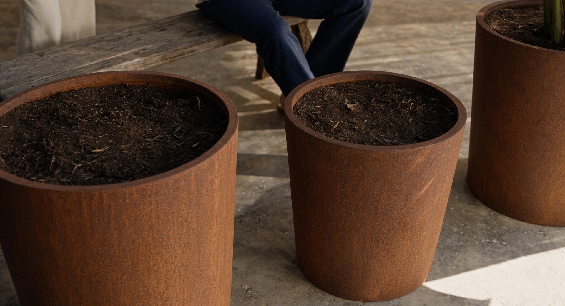 Pots de fleurs ronds élancés Clio en acier Corten remplis de terreau, sur un sol en béton près d'un banc