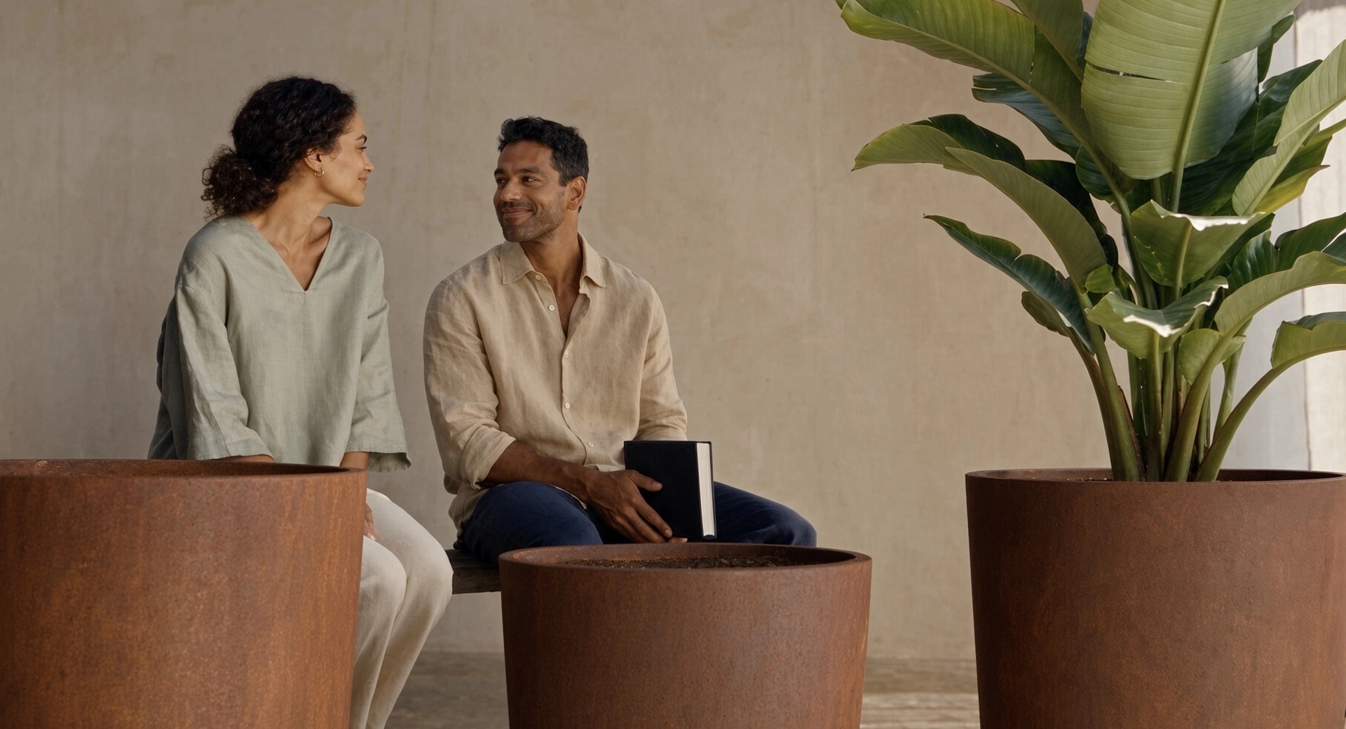 Pots de fleurs ronds élancés Clio en acier Corten, l'un garni d'une plante à larges feuilles, près d'un couple sur un banc