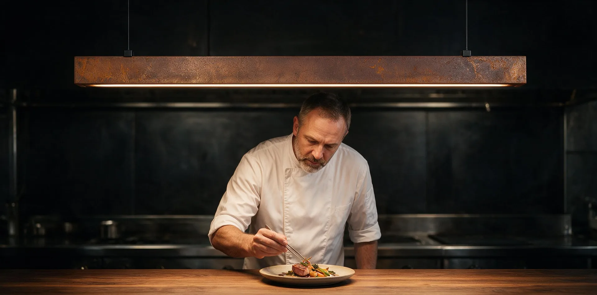 Suspension Lyra en acier Corten éclairant un chef qui dresse une assiette sur un comptoir en bois, cuisine sombre