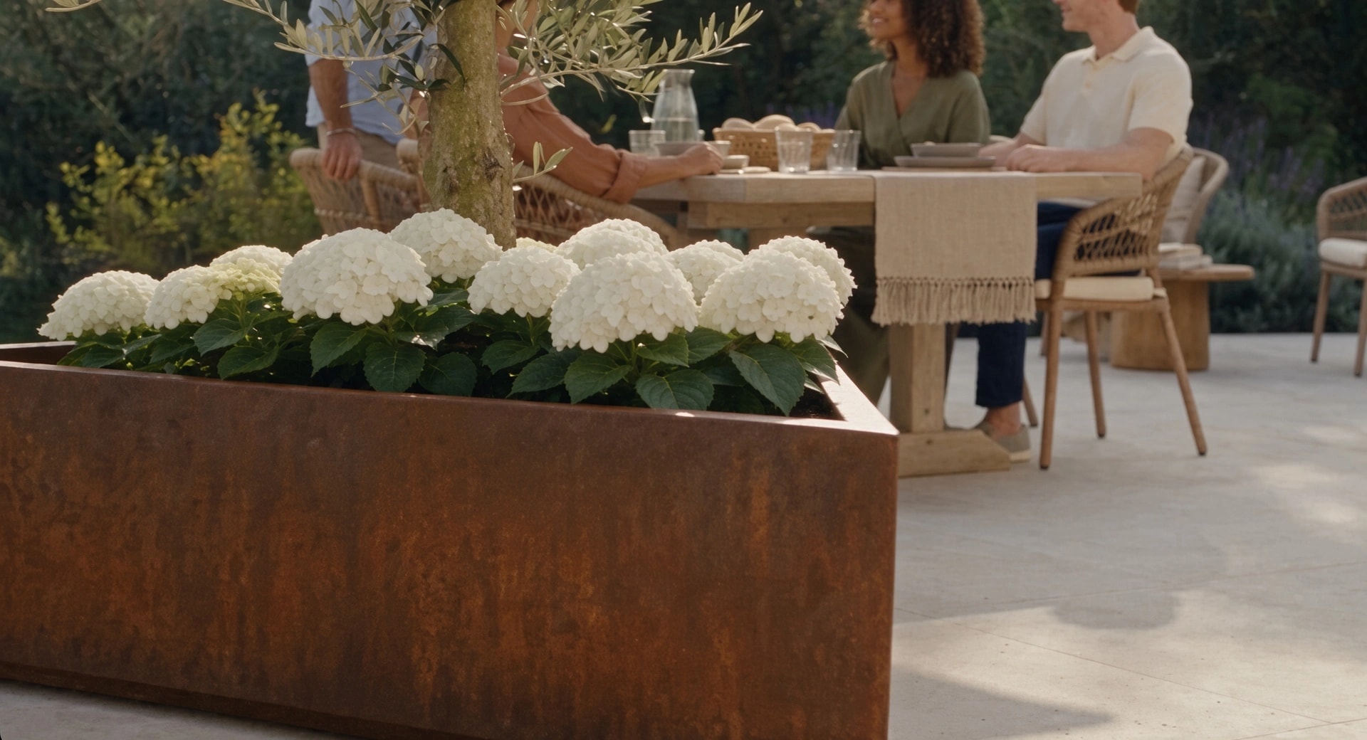 Jardinière triangulaire Iris en acier Corten aux hortensias blancs au premier plan, des convives attablés derrière