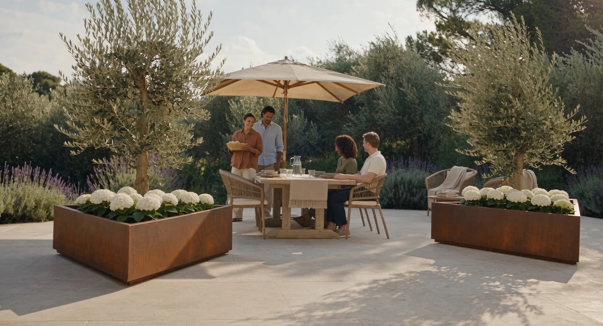 Deux jardinières triangulaires Iris en acier Corten plantées d'oliviers, encadrant une table de jardin sous parasol