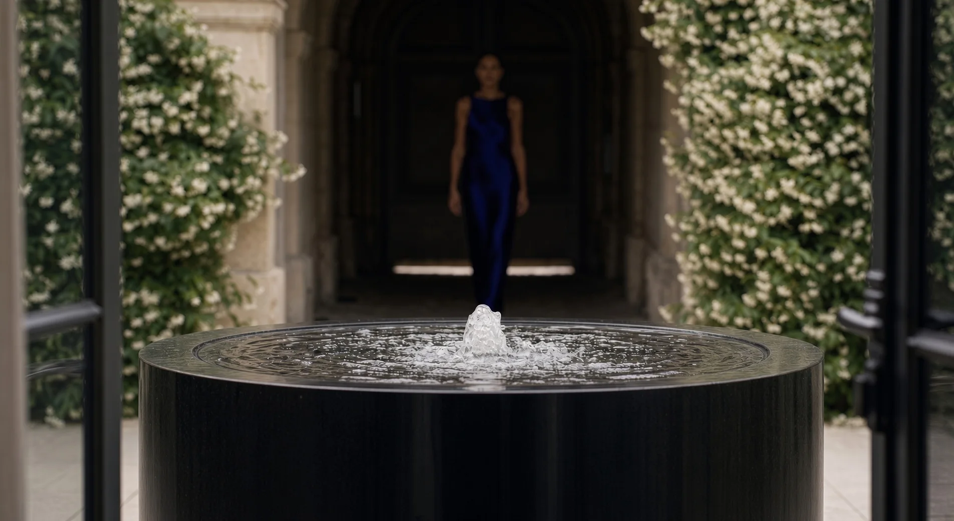 Fontaine ronde Elio en aluminium noir cadrée entre deux grilles ouvertes, jet bouillonnant au premier plan