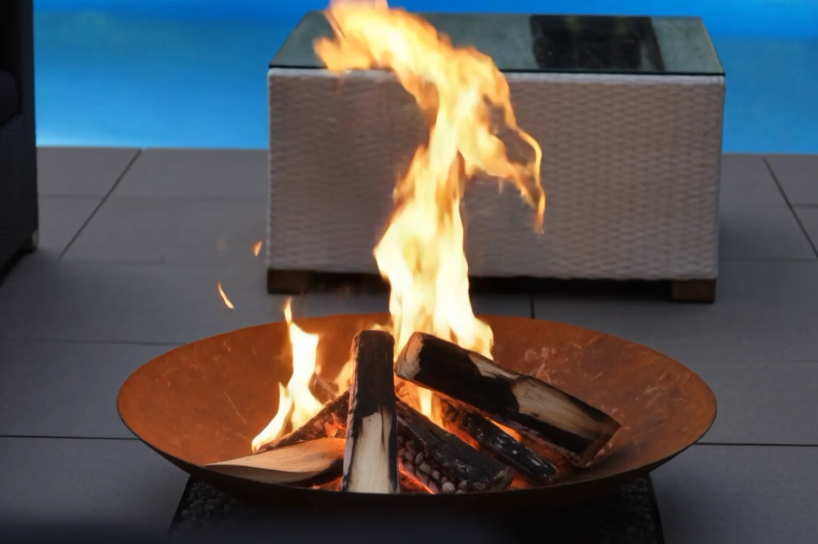 Bol de feu Vesta en acier Corten allumé devant une table basse en résine tressée blanche, piscine bleue au fond