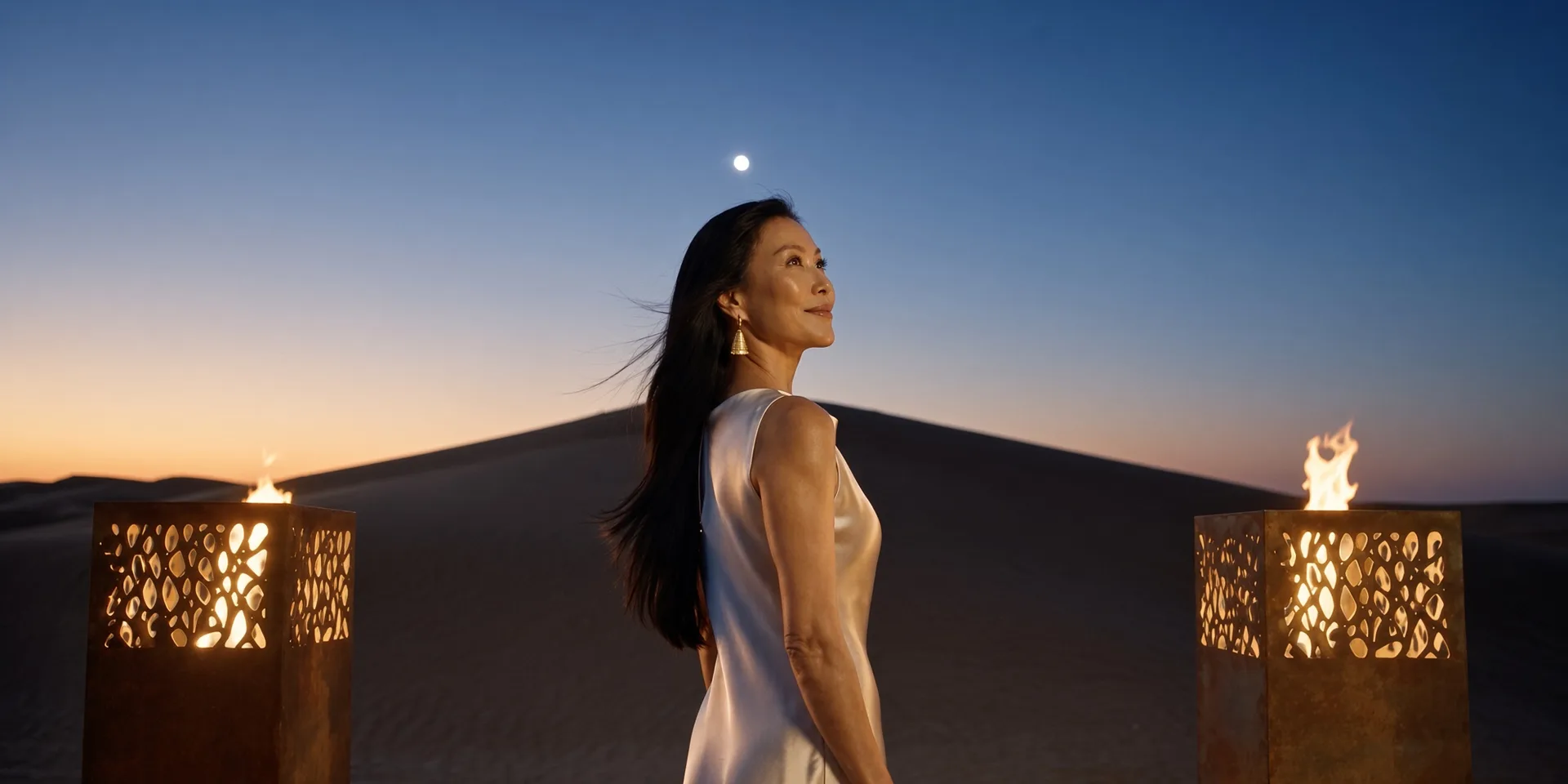 Une femme entre deux lanternes Vesper en acier Corten allumées, dunes et lune au crépuscule