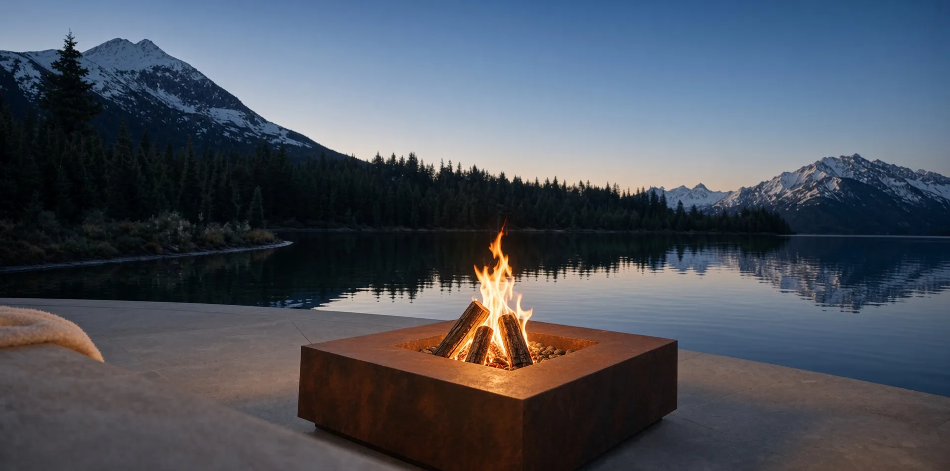 Table de feu Héphaïstos en acier Corten allumée au bord d'un lac de montagne, au crépuscule