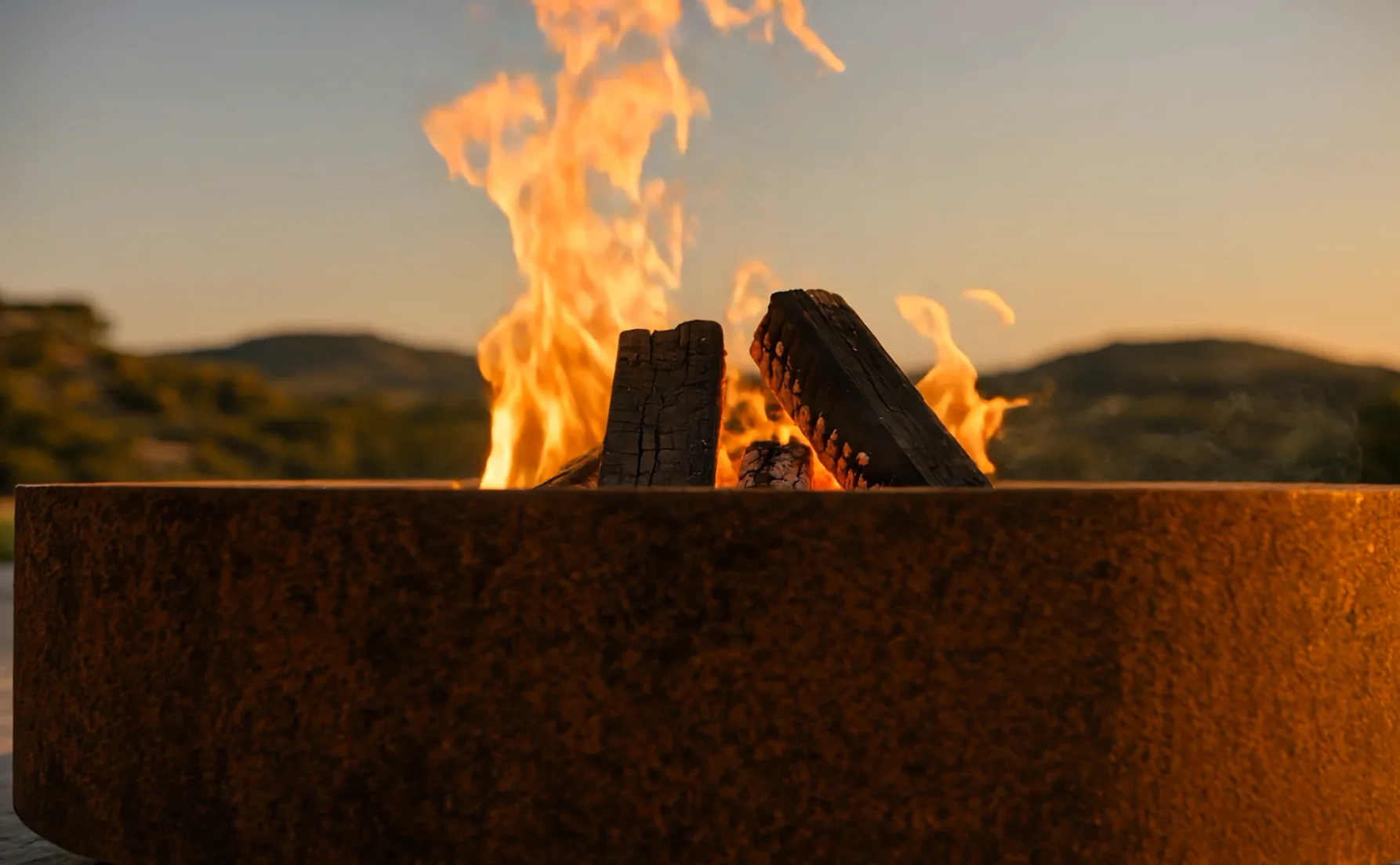 Nahaufnahme des dicken Rands des Feuertischs Hélios aus Cortenstahl, Flammen und Scheite im Sonnenuntergang