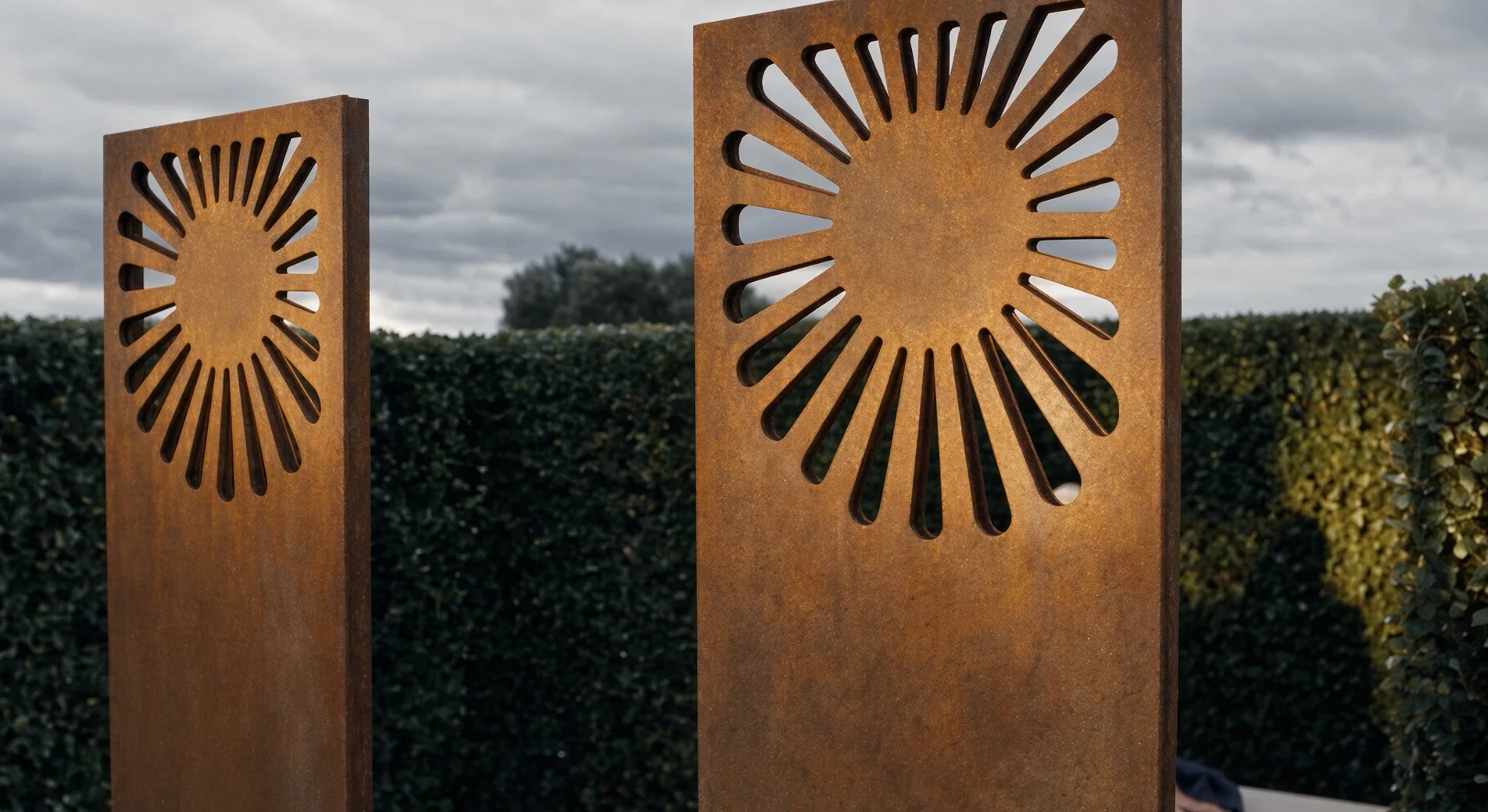 Soleils ajourés de deux panneaux décoratifs Éole en acier Corten, vue en contre-plongée sous un ciel nuageux