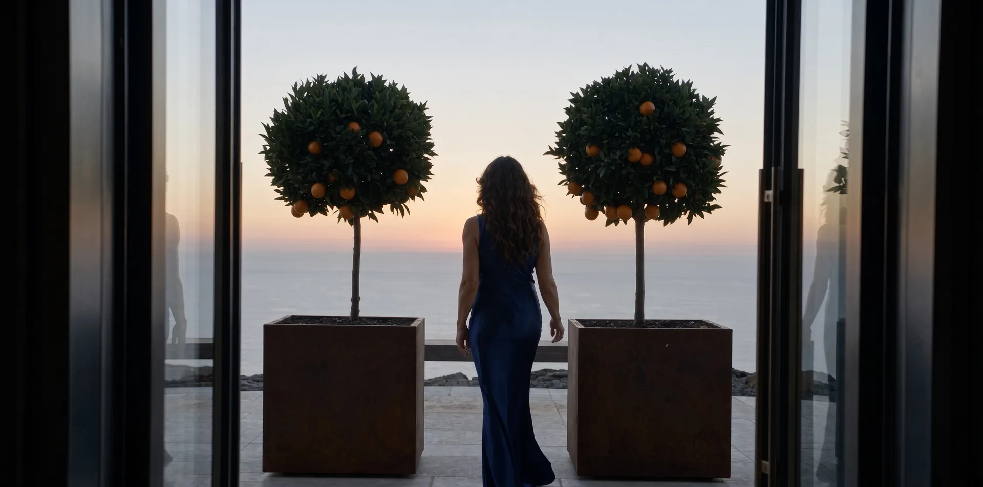 Jardinières carrées Cybèle en acier Corten plantées d'orangers, une femme entre elles face à la mer, vue d'une baie vitrée