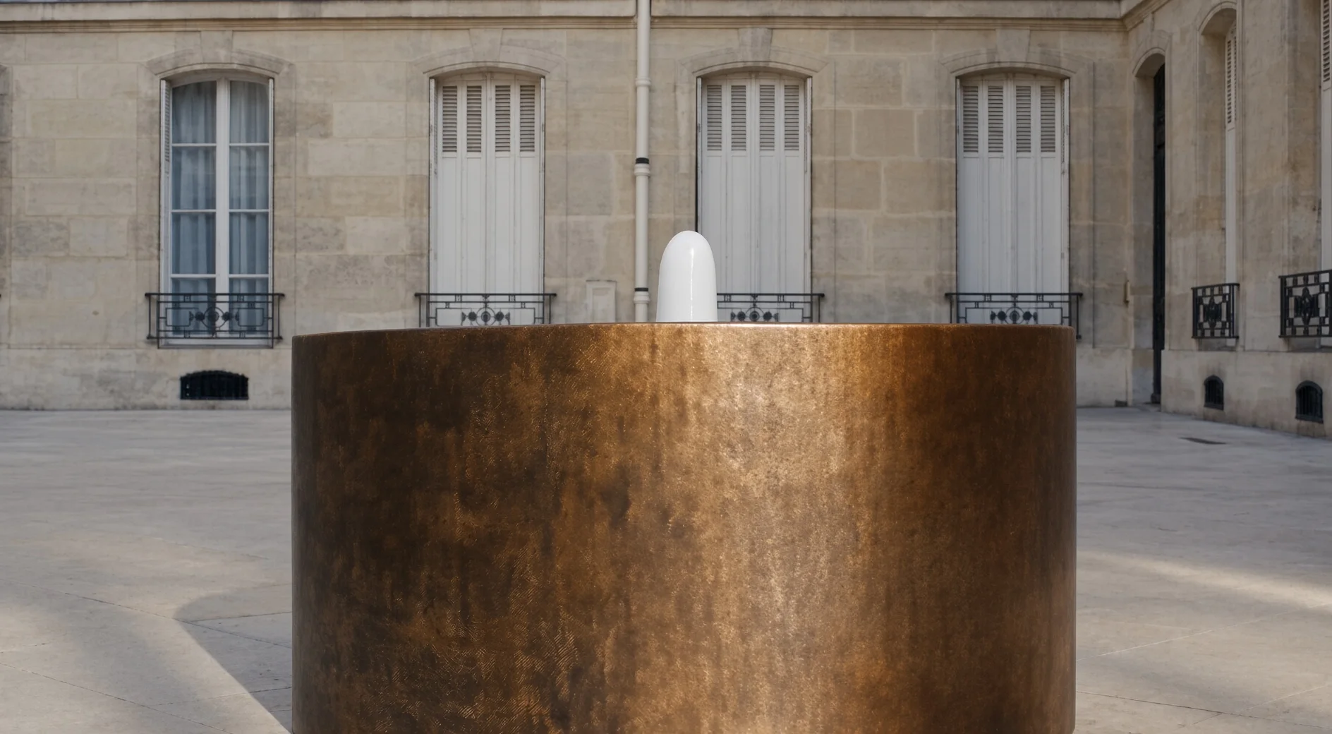 Fontaine ronde Elio en acier Corten en cadrage bas, façade en pierre de taille à hautes fenêtres en arrière-plan