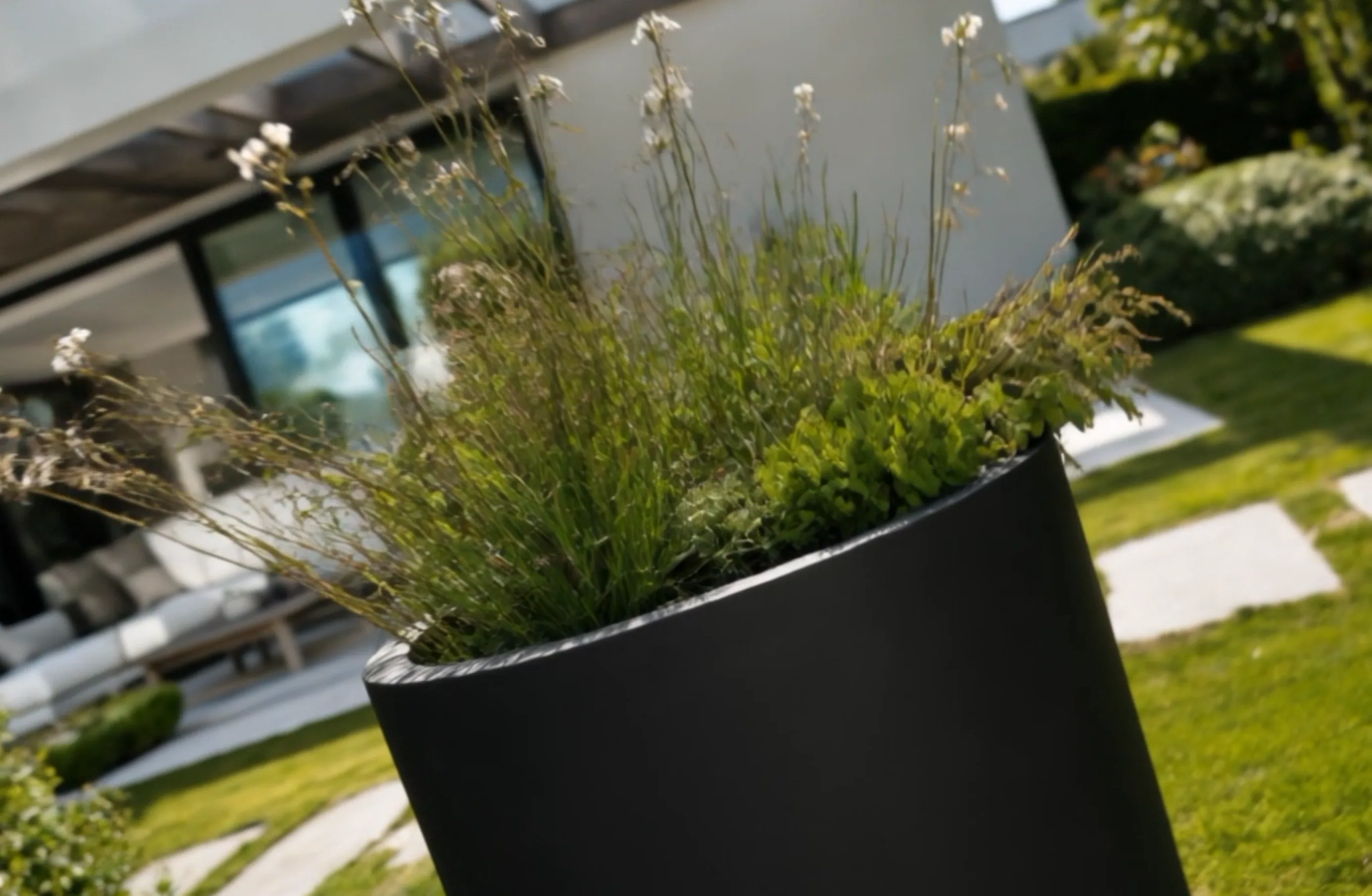 Pot de fleurs rond Callisto en aluminium noir et ses graminées en cadrage incliné, maison floue au fond