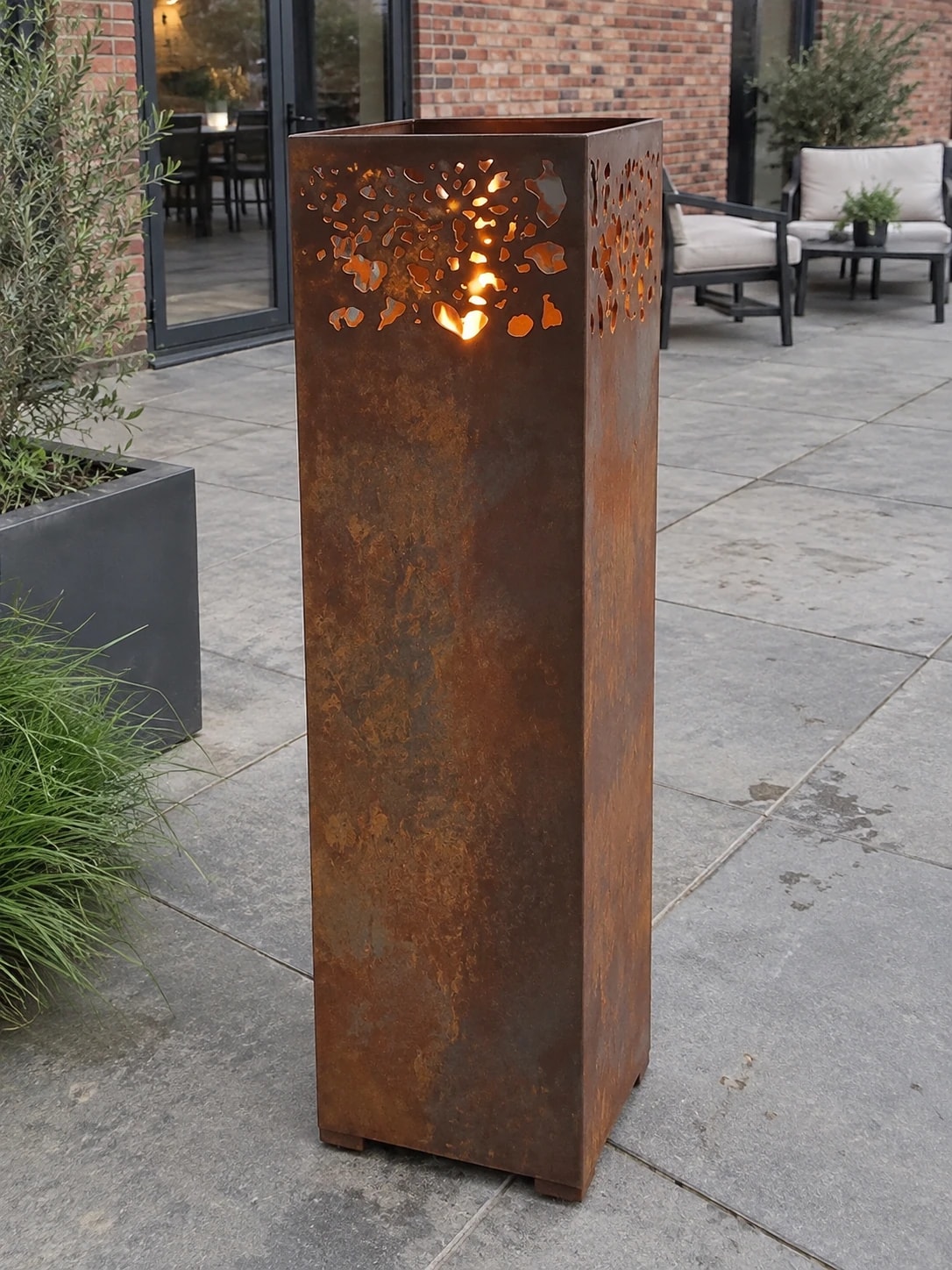 Lanterne Aurore en acier Corten allumée sur une terrasse dallée devant un bâtiment en brique, salon d'extérieur