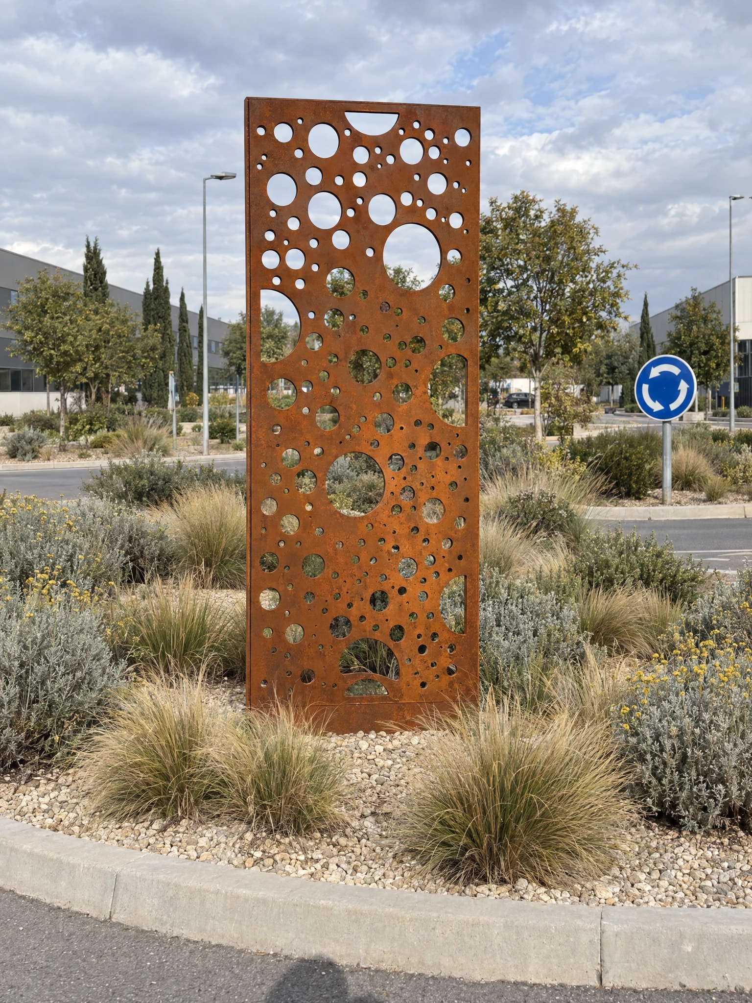 Panneau décoratif Notos en acier Corten dressé sur un rond-point planté de graminées, sous un ciel nuageux
