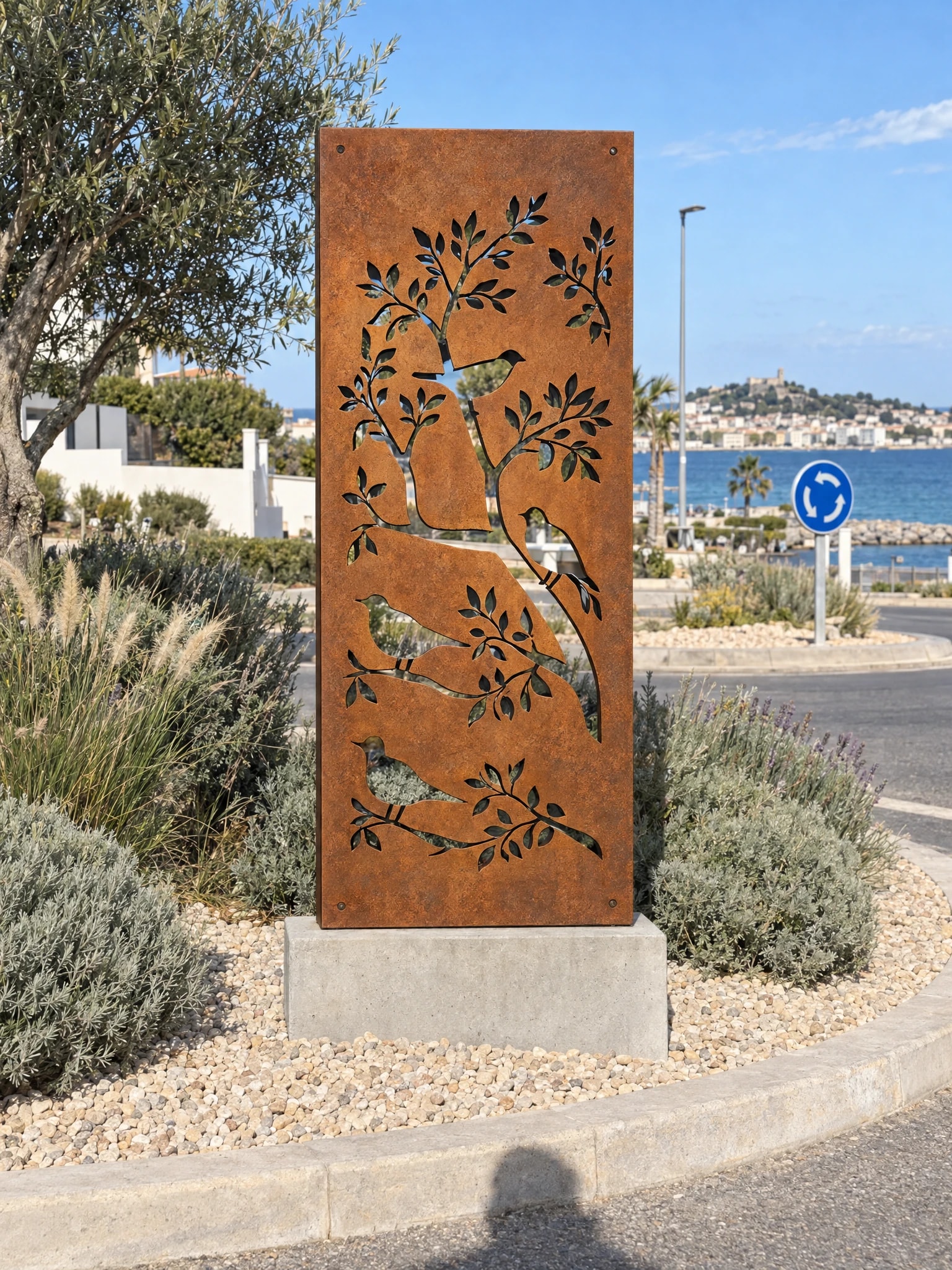 Panneau décoratif Sirocco en acier Corten sur un socle au centre d'un rond-point planté, mer en arrière-plan