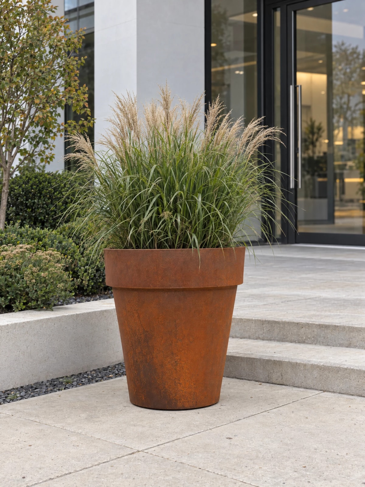 Vaso conico Calypso in acciaio Corten con graminacee, ai piedi dei gradini di un ingresso vetrato