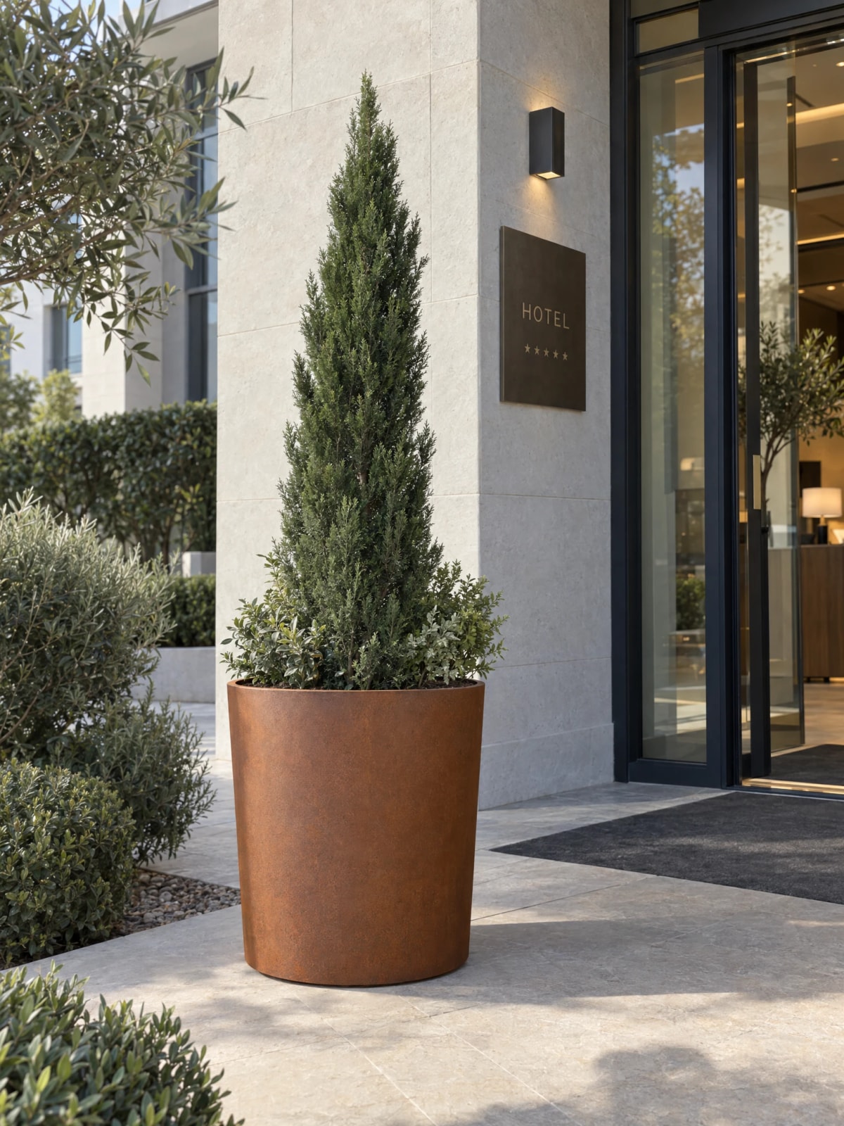 Vaso rotondo slanciato Clio in acciaio Corten con una conifera a cono all’ingresso di un hotel
