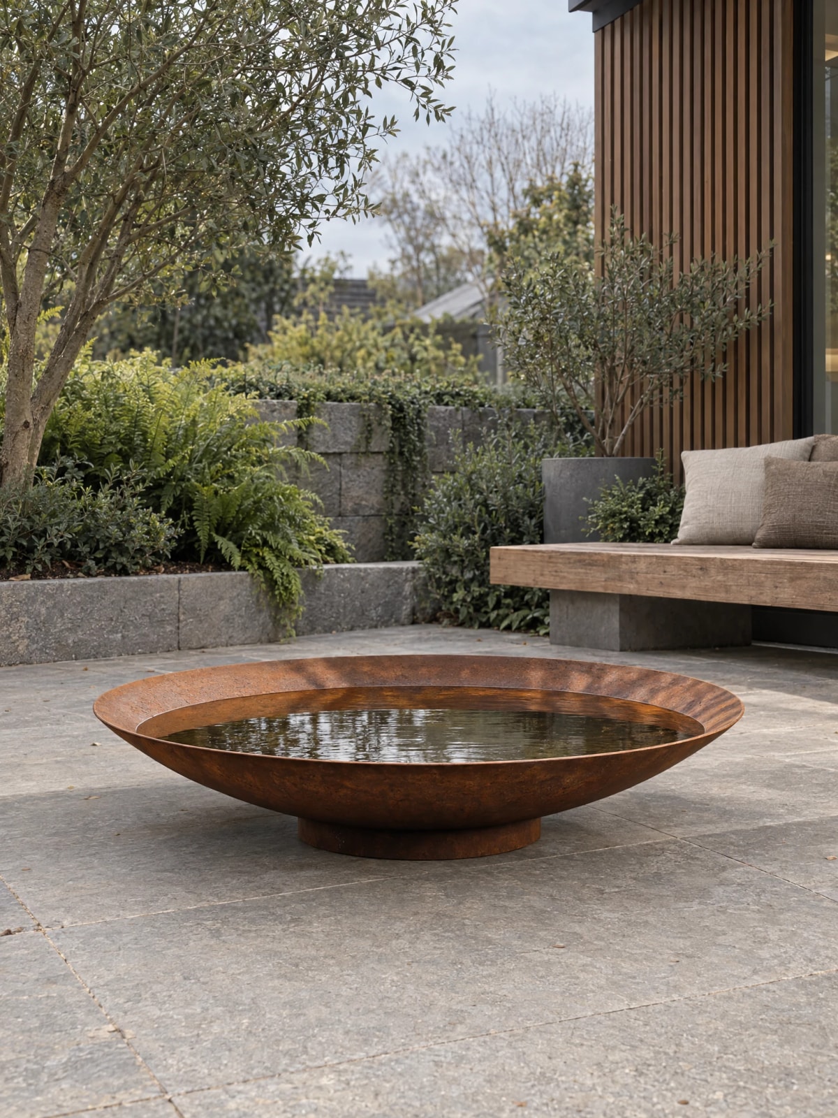 Wasserschale Sélène aus Cortenstahl vor einem Infinity-Pool und einer Outdoor-Lounge, Meerblick in der Dämmerung