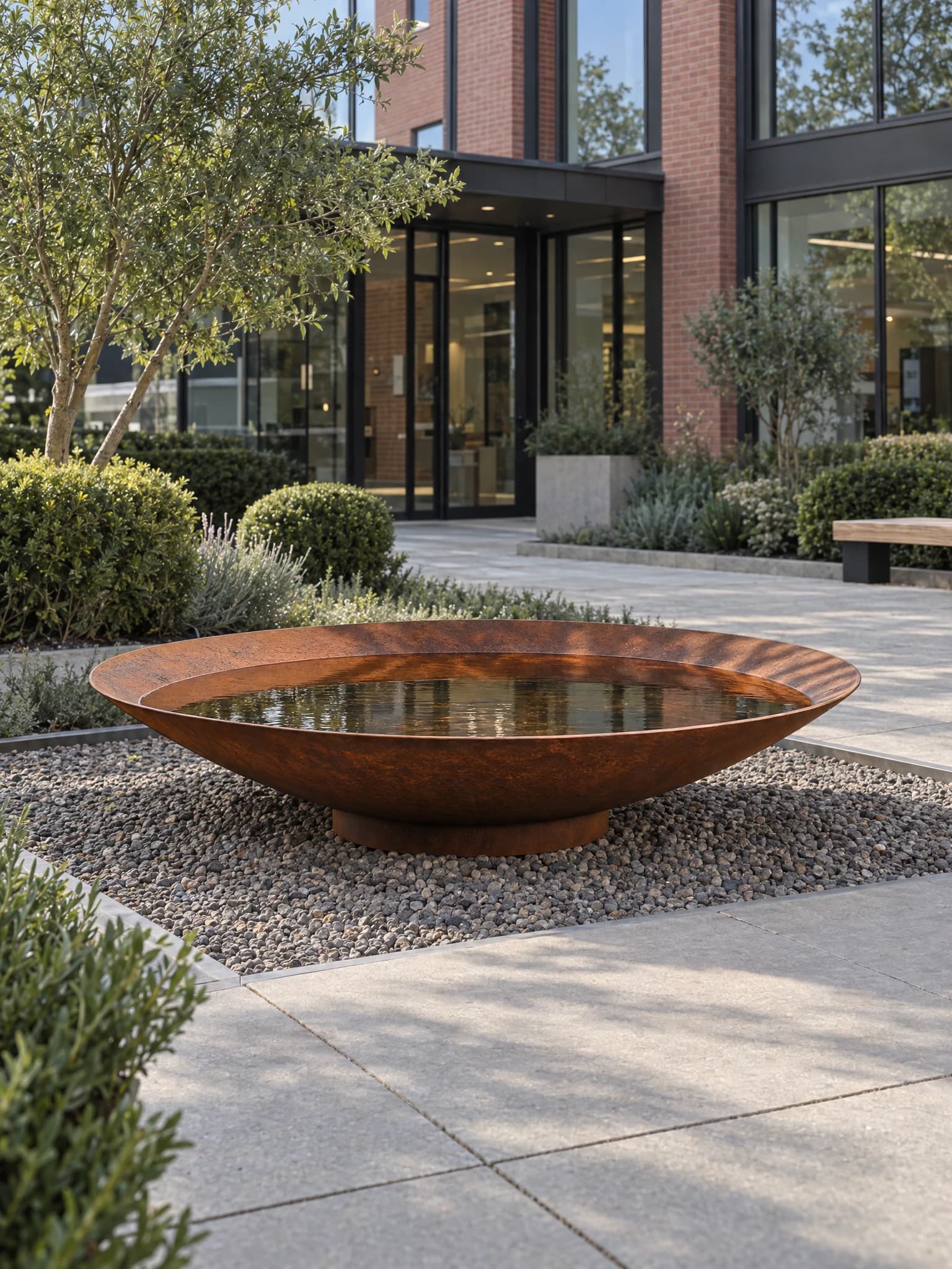 Vasque d'eau Sélène en acier Corten, large coupe basse remplie d'eau sur un lit de gravier devant un bâtiment contemporain